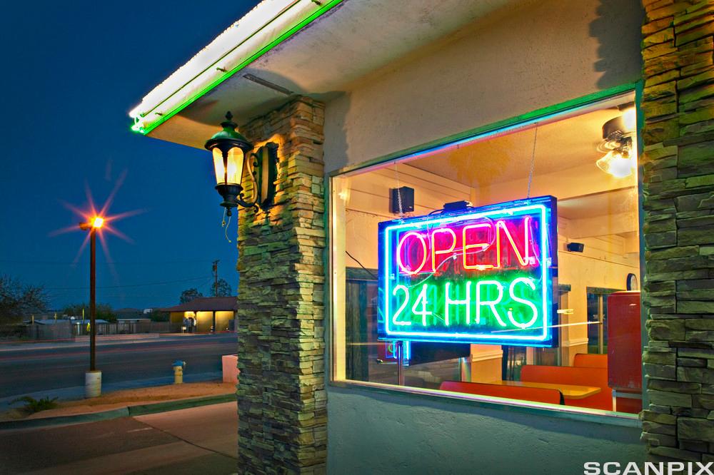 Eit neonskilt med skrifta "Open 24 H R S" heng i vindauget på ein kafé. Det er kveld og ingen menneske verken utanfor eller i kaféen. Foto.