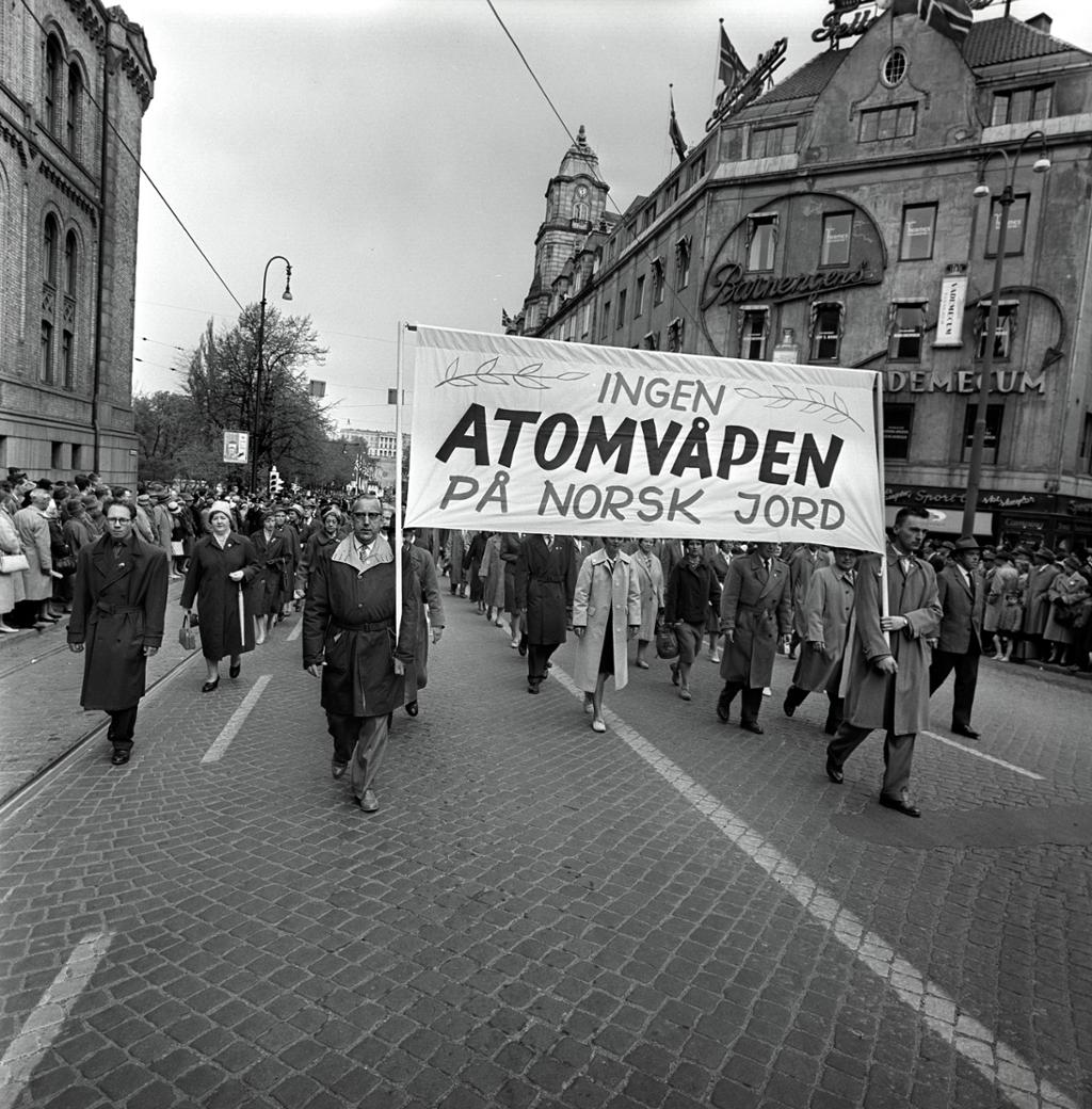 Demonstrasjonstog med mange mennesker både i toget og ved siden av. To menn holder et banner med teksten "Ingen atomvåpen på norsk jord". Svart-hvitt foto.