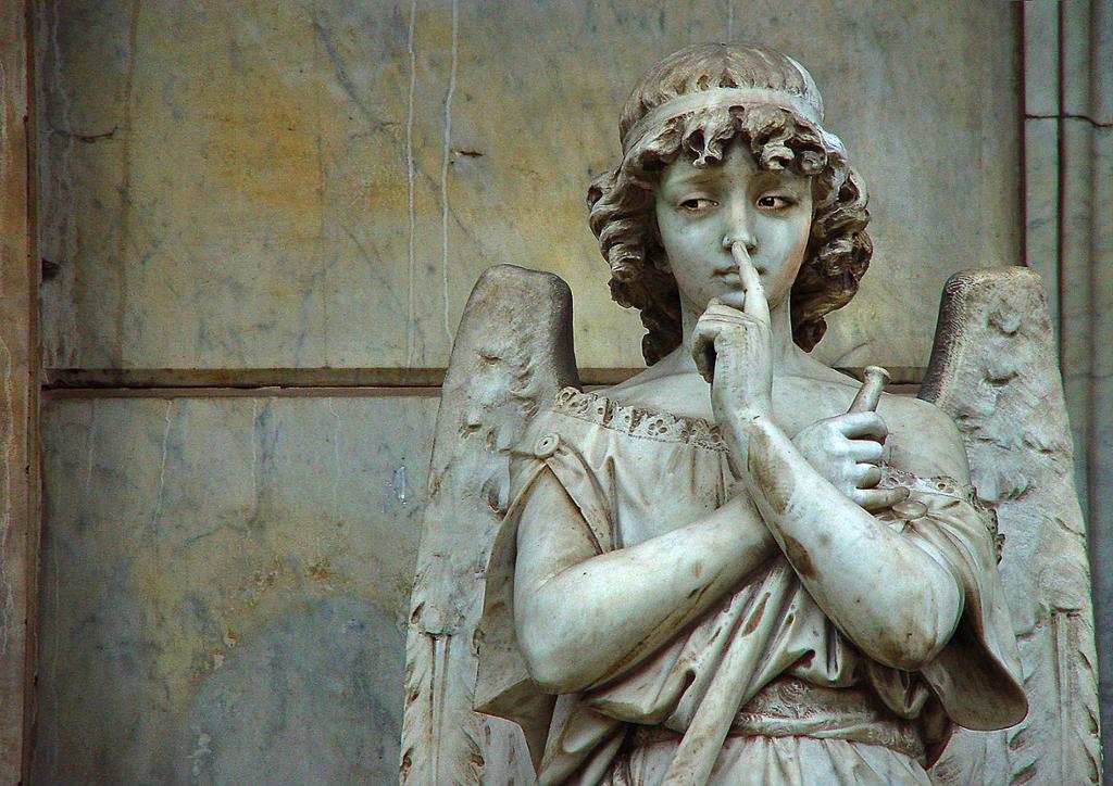 Ein statue av ein engel. Engelen ser til sides og held den eine fingeren mot nasen. Han har kjole, panneband, krøllete hår og venger. Foto.