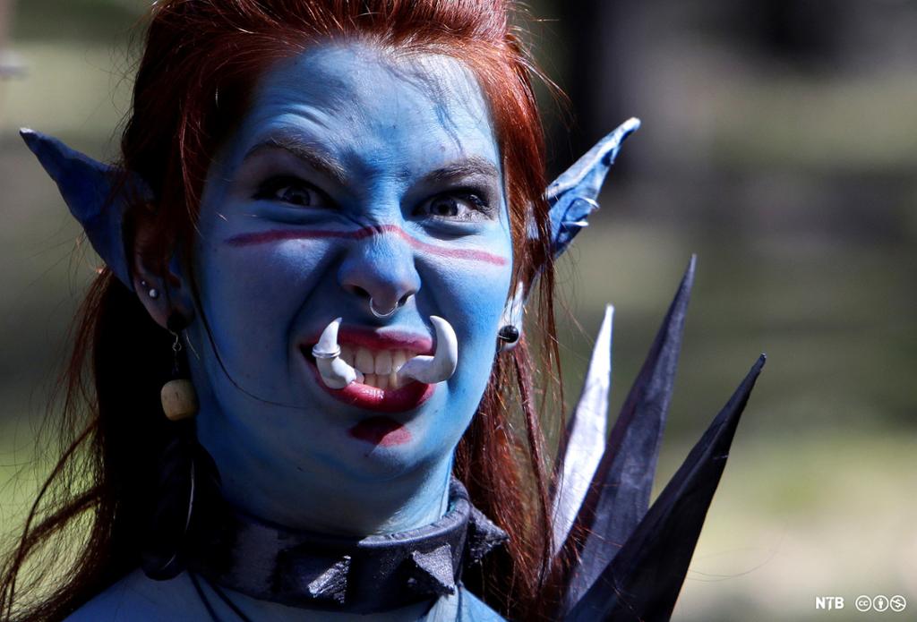 Ung kvinne med blå maling i ansiktet og lange, spisse ører, ring i nesen og spesielle tenner som står opp fra munnen. Hun har et bånd med store nagler rundt halsen. Foto. 