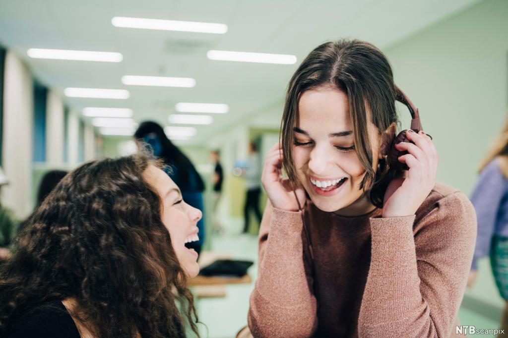 To jenter hører på musikk i hodetelefoner og ler i en korridor. Foto.