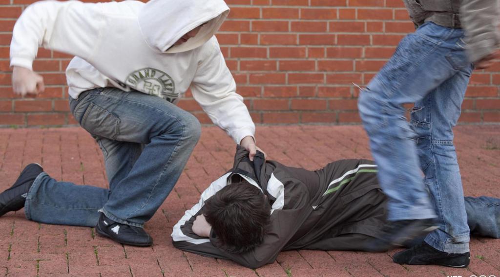 En gutt ligger på bakken mens han blir slått og sparket av to andre. Foto.