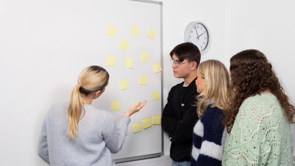 Fire elever står foran ei tusjtavle med post-it-lapper. En av dem forklarer noe til de andre. Foto. 