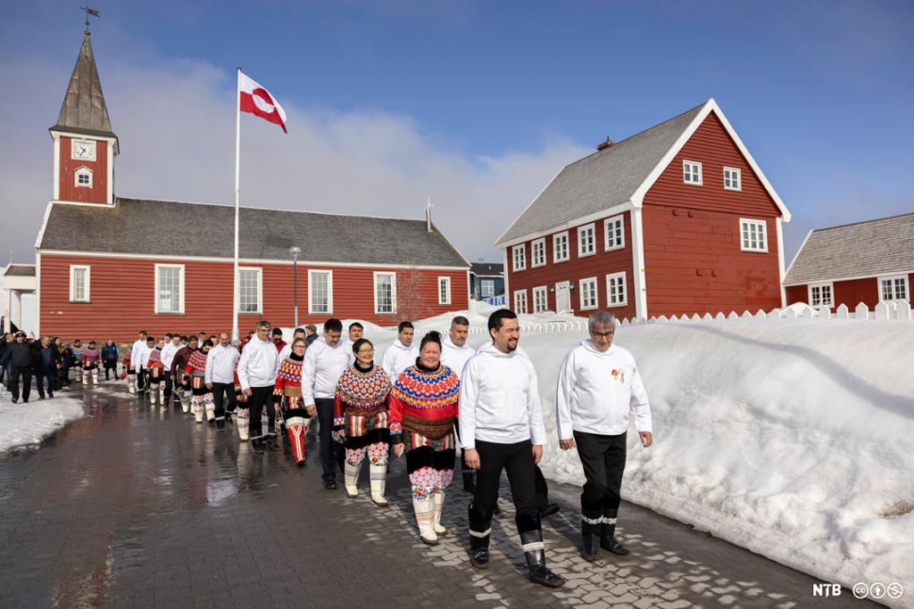 Medlemmer av parlamentet på Grønland i klassiske drakter går frå ei raud trekyrkje. Foto. 