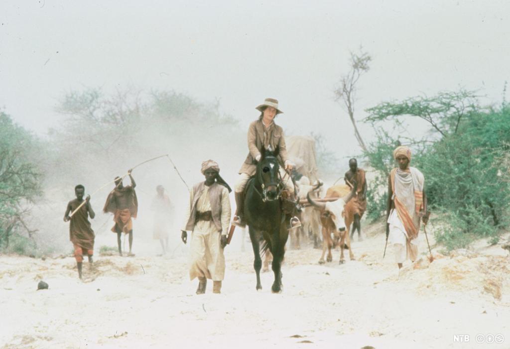 Skuespilleren Meryl Streep i kakiklær sitter til hest omgitt av afrikanske gjetere. Foto fra filmen "Out of Africa".