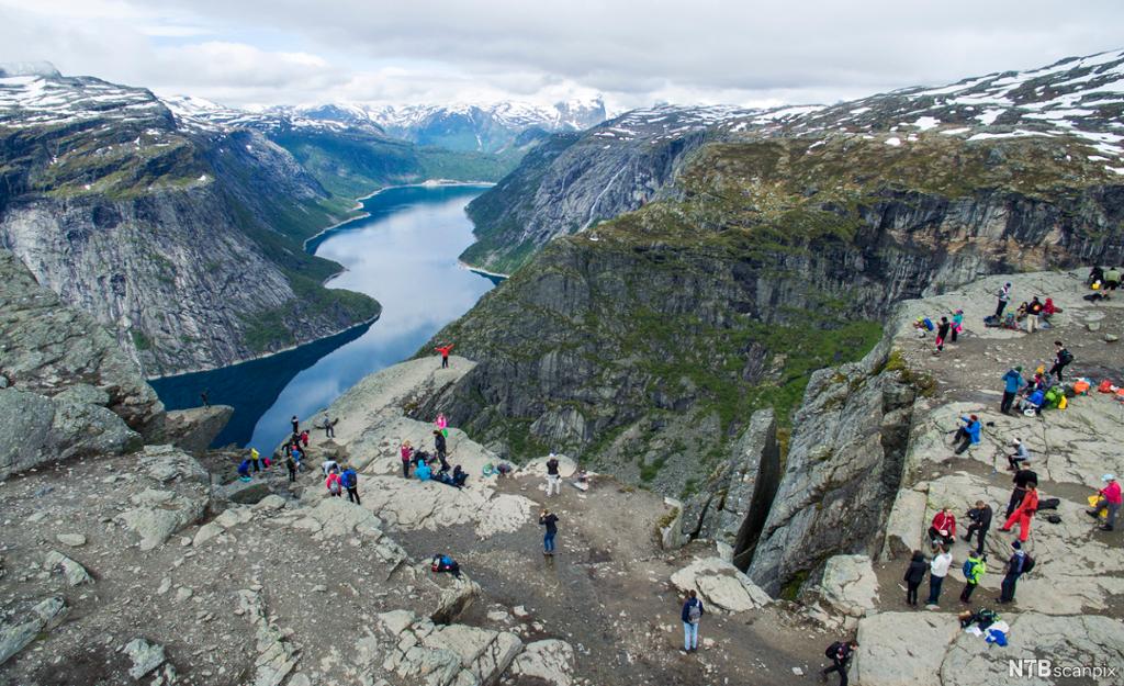 Mange personar på eit fjell med utsikt til ein fjord og snøkledde fjell. Foto.