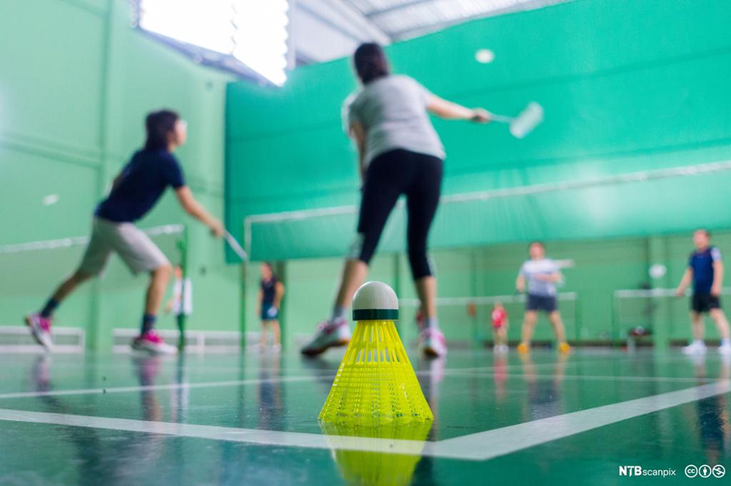 Fjørball på gulv i forgrunnen, personer som spiller badminton i bakgrunnen. Foto.