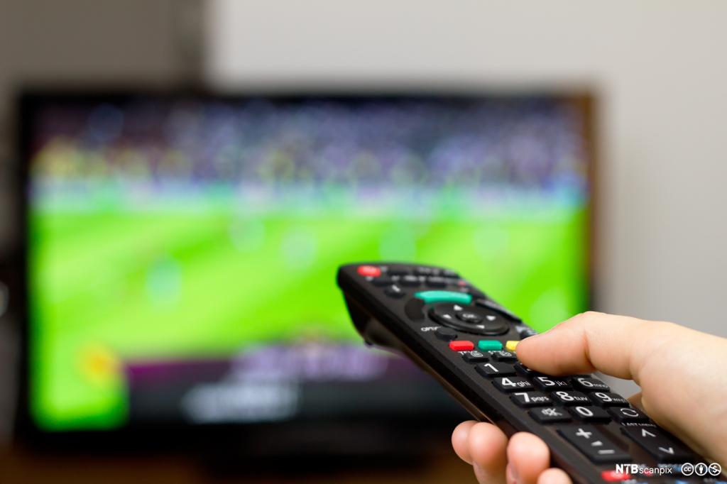 Hånd med TV-kontroll foran sportssending. Bilde.