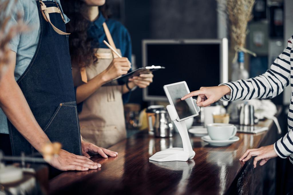 Tre personer står rundt en digital bestillingsskjerm på en selvbetjent kaffebar. Foto.