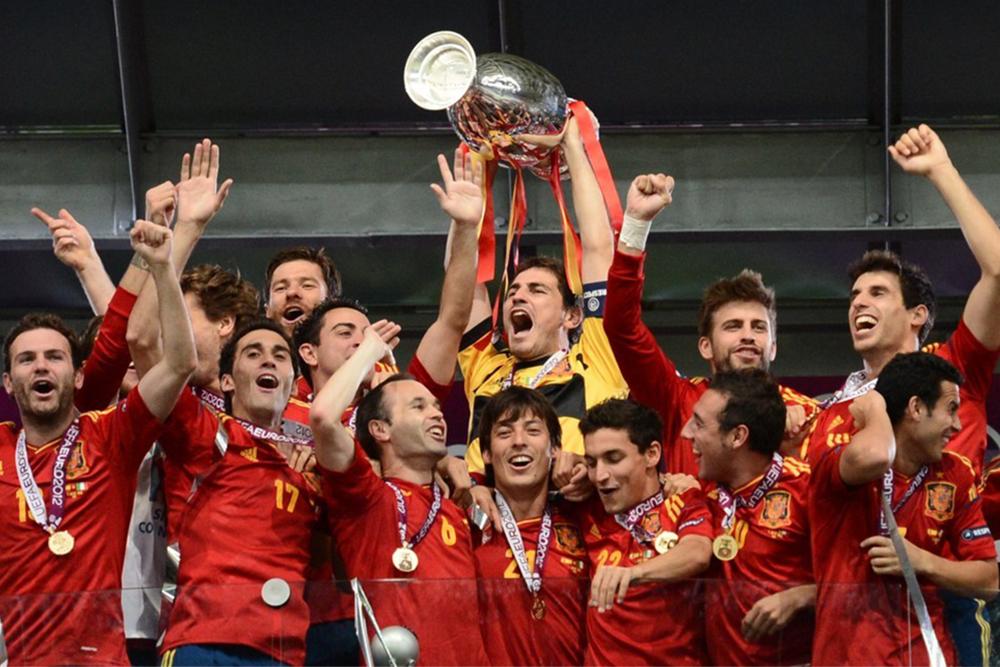 Photo: A football team (the Spanish national team) are celebrating a victory. They wear medals and one player (the keeper) is holding a cup aloft. 