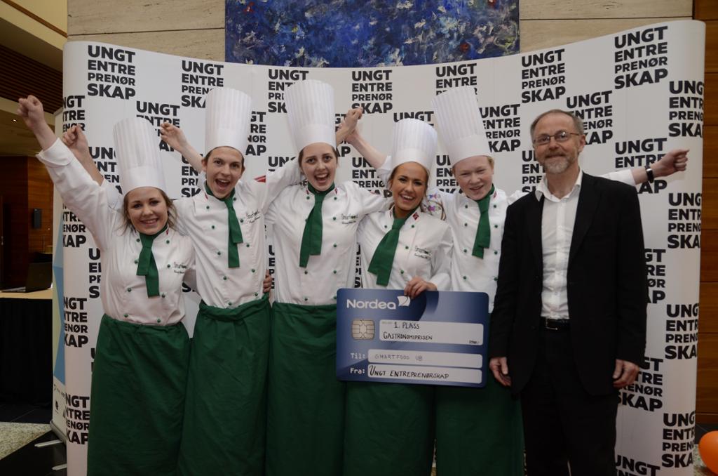Fem blide unge kokker strekker hendene i været og jubler. Den ene holder en plakat utformet som et bankkort hvor det står 1. plass Gastronomiprisen til Smart food UB fra Ungt entreprenørskap. En smilende mann i dress står ved siden av dem. Foto.
