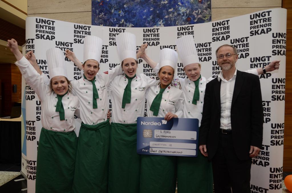 Fem blide unge kokker strekker hendene i været og jubler. Den ene holder en plakat utformet som et bankkort hvor det står 1. plass Gastronomiprisen til Smart food UB fra Ungt entreprenørskap. En smilende mann i dress står ved siden av dem.  Foto. 