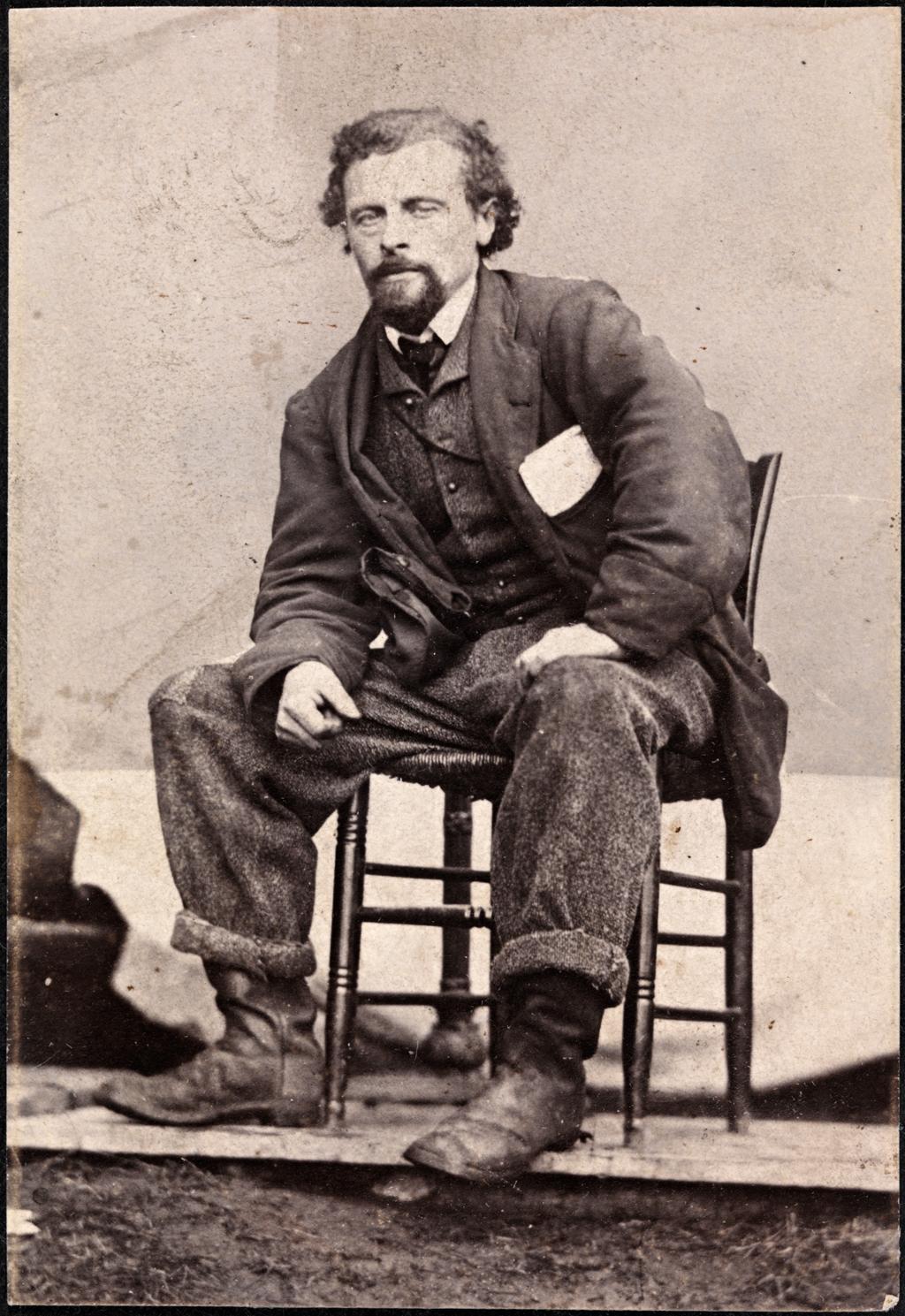 Portrett av Marcus Thrane ca. 1863. Han sitter bredbeint og litt framoverlent på en stol. Han er uvørent kledd, buksa er oppbretta og har lapp på kneet. Han har noen papirer i brystlomma. Foto. 