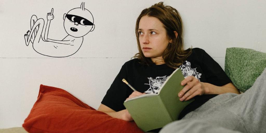 Ung kvinne ligger og lener seg mot en vegg mens hun skriver i ei bok. På veggen bak henne er det en naivistisk tegning av en naken person med en svart maske over øynene. Foto.