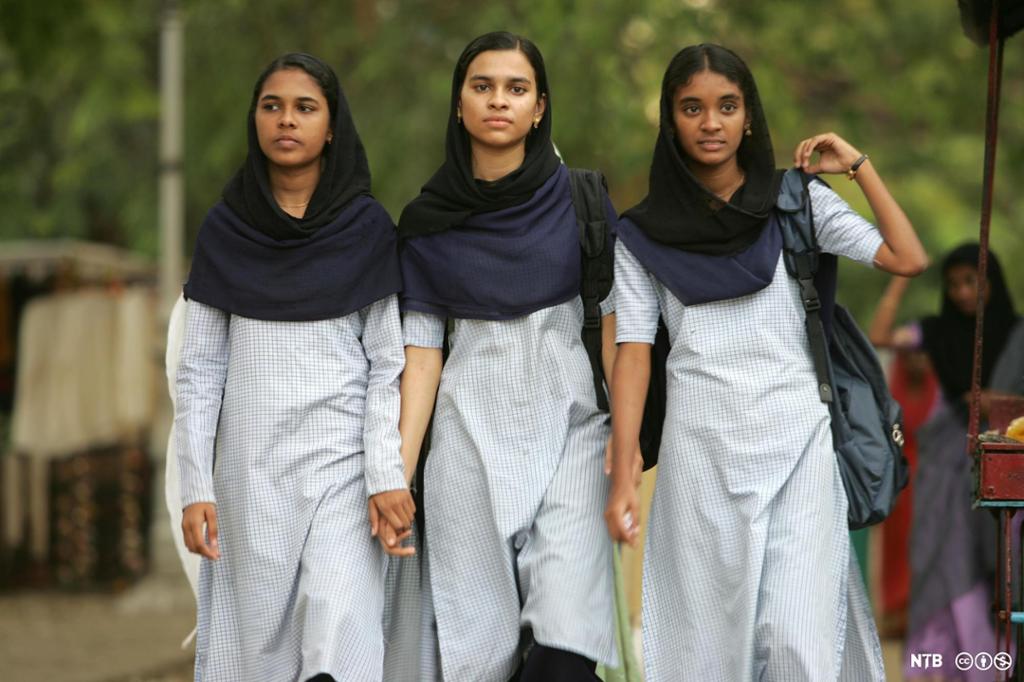 Three Indian girls walking hand in hand, They are dressed in similar saris or school uniforms. They look serious and focused. 