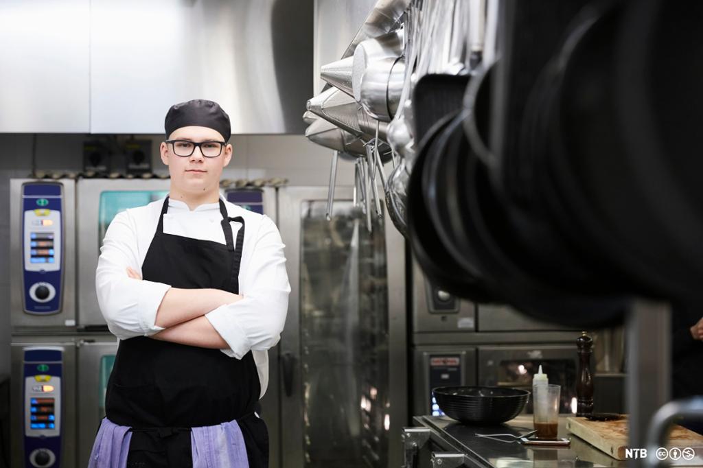 Ein ung kokk med armane i kross står framfor omnane på eit kjøkken. Foto.