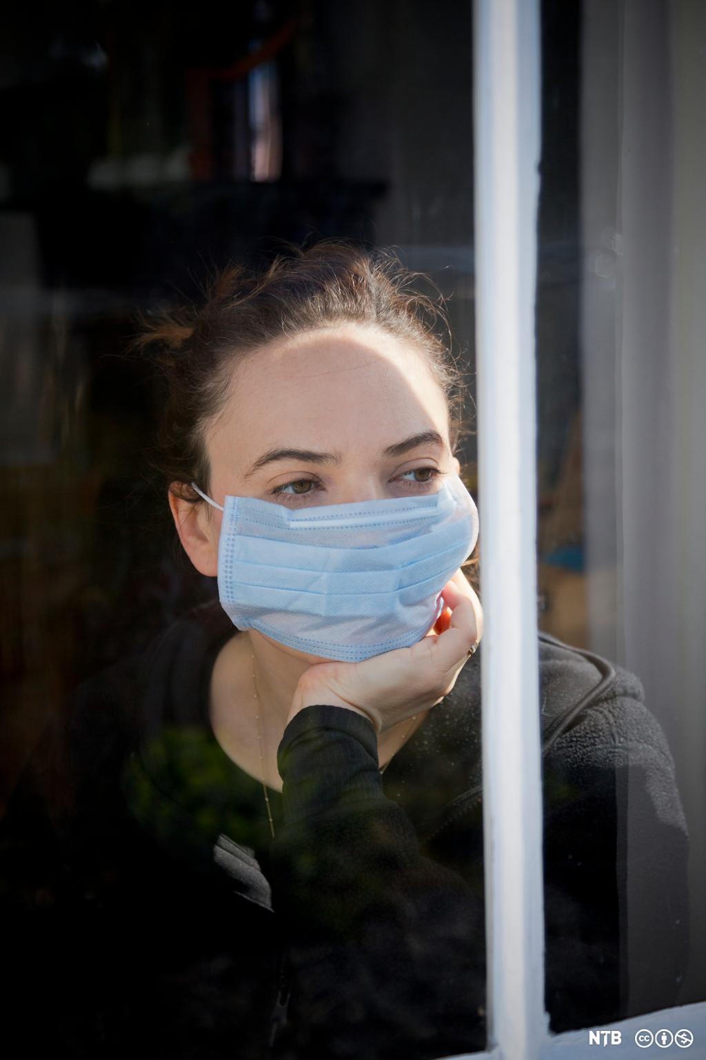 Kvinne har på seg et munnbind og sitter og ser ut av vinduet. Foto.
