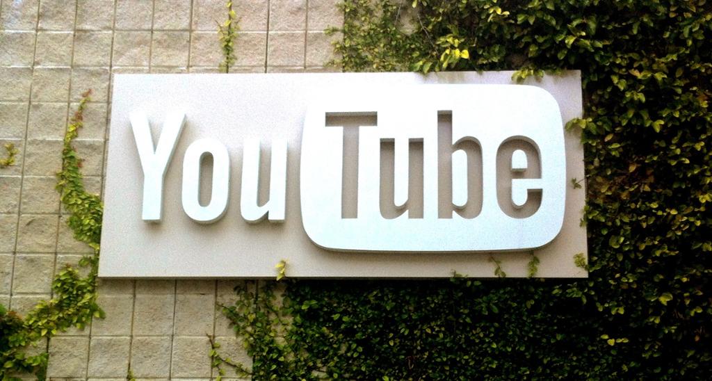 Et hvitt YouTube-skilt som henger på en vegg med eføy. "You" er med uthevede bokstaver, "Tube" er stanset ut fra ei plate med avrunda hjørner. Foto.