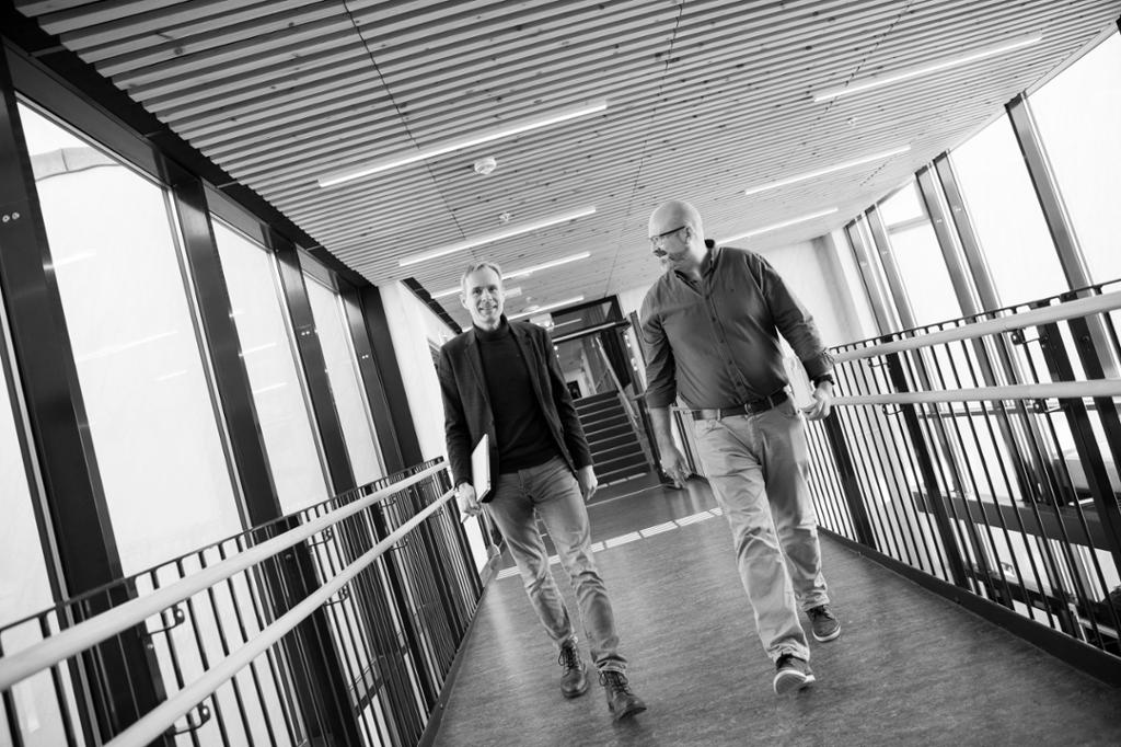 Sort hvitt foto, noe skråstilt. Motiv: Innendørs, to menn i helfigur, gående mot kamera.
