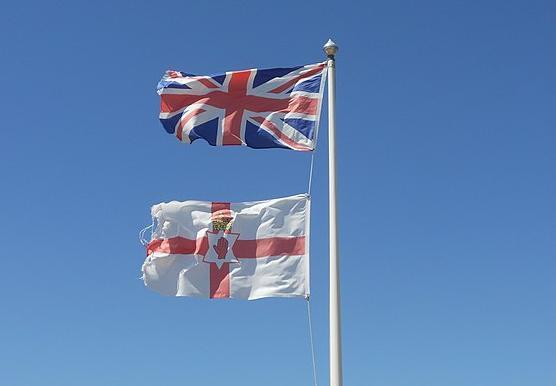 A flagpost with two flags: the Union Jack on the top and the Ulster Banner at the bottom. The Ulster banner has a red cross on a white background, showing the palm of a red hand in the middle of the flag. Photo.