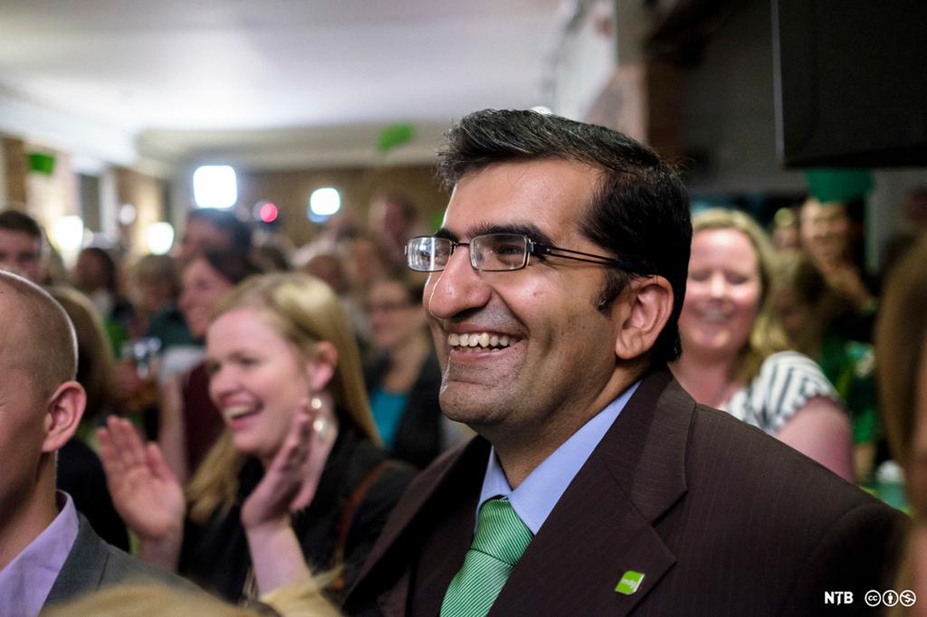 En mann i dress, skjorte og slips og med grønn pin på skjortekragen smiler. I bakgrunnen er det flere personer som smiler. De sitter alle i et forsamlingslokale. Foto.