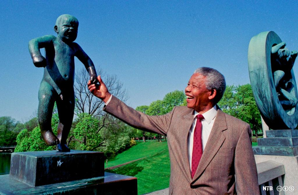 Photo: Nelson Mandela holds the hand of Gustav Vigeland's sculpture 'Sinnataggen' in Vigelandsparken in Oslo. He is smiling. He is wearing a brown suit and a red tie. 