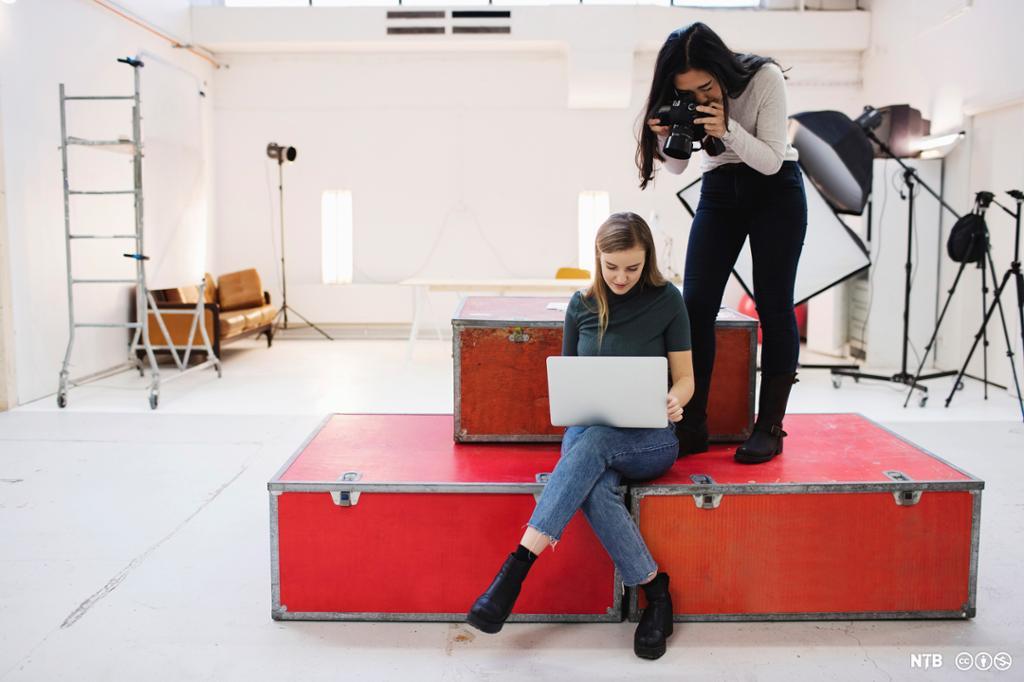 I et studio sitter en jente på en stor rød flykasse med en laptop i fanget. En annen jente står bak henne og fotograferer skrått over skulderen hennes. Foto.