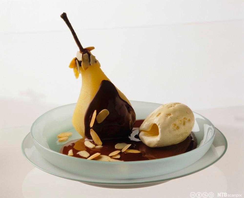 Posjert pære i en skål, pent anrettet med vaniljeis, sjokoladesaus og mandelflak. Foto.