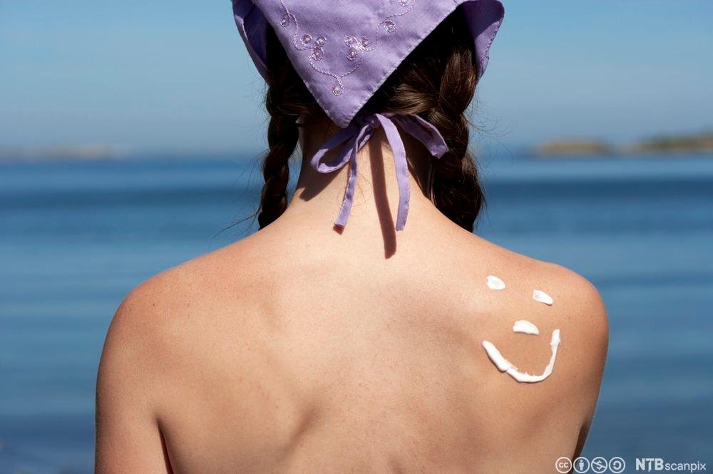 Jente som soler seg. Noen har brukt solkrem til å tegne et smilefjes på ryggen hennes. Foto.