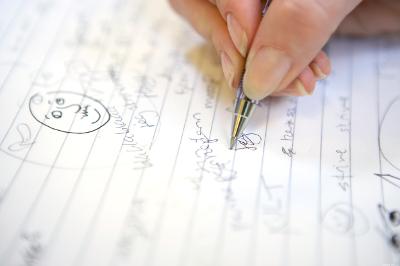 Photo: Person writing in an open notebook. The open page has scribbled drawings and notes. 