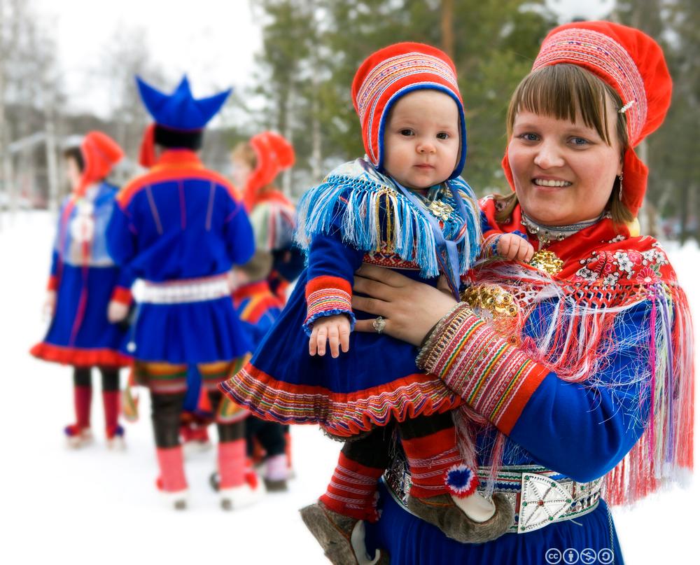 Samisk kvinne bær eit lite barn. Begge er kledde i samekufter. Foto.