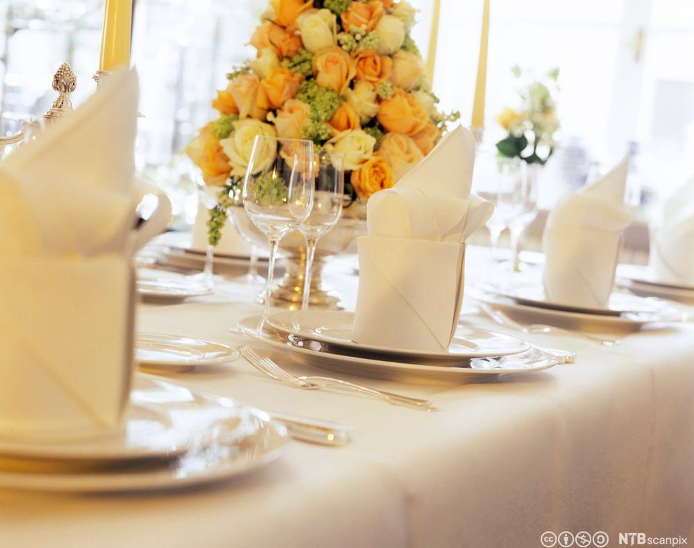 Eit pent dekt bord med kvit duk, kvite tallerkenar, vinglas, og store blomsteroppsatsar. Foto.