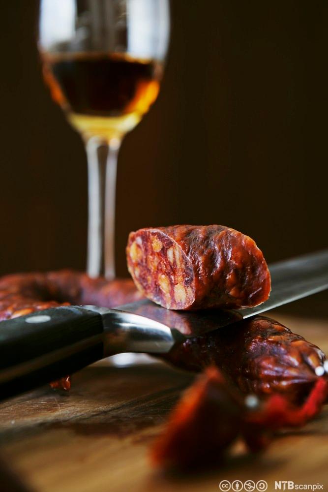 Bilde av en chorizopølse på en kniv, og et glass sherry. Foto.