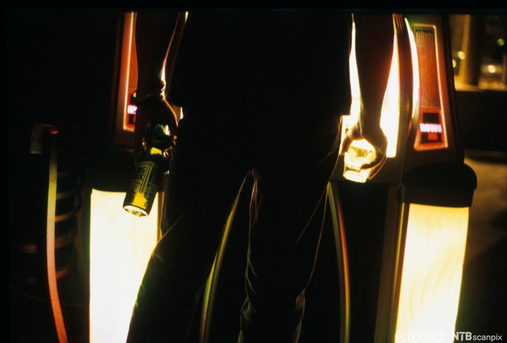 Mørkt stemningsbilde av en ung mann på bar. Han står med ryggen til og har en ølflaske i den ene hånda og et glass i den andre. Foto.ung mann på bar