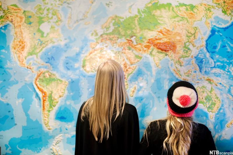 To elever står med ryggen til og betrakter et stort verdenskart på en vegg. Foto.