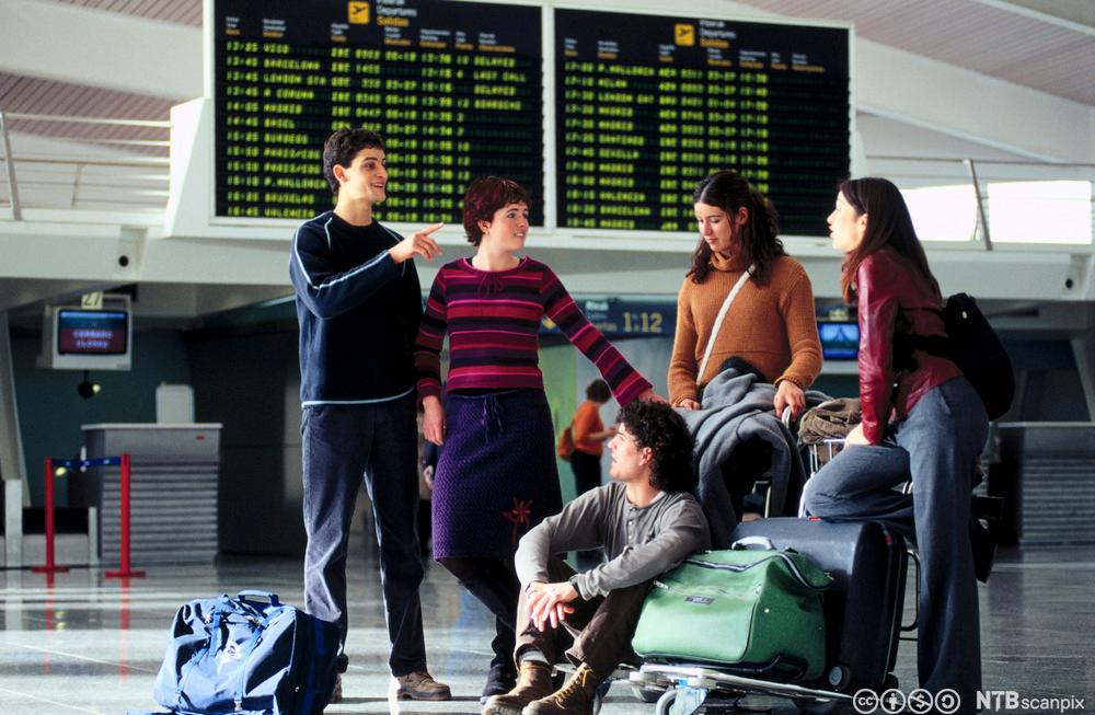 Fem ungdommar på ein flyplass. Dei har med seg bagasje, og noko av bagasjen er plassert på ei bagasjetralle. Foto.
