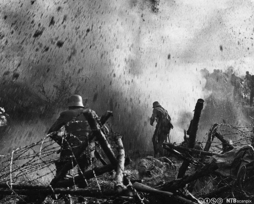 Soldater står på slagmarken i bomberegn med piggtrådstolper rundt seg. Foto.