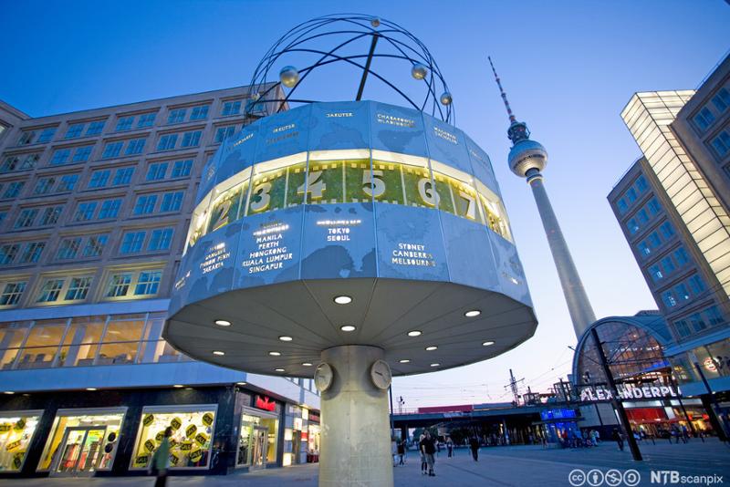 Bilde av Alexanderplatz i Berlin om kvelden. I forgrunnen et byggverk i betong som viser hva klokka er i ulike byer rundt om i verden. I bakgrunnen står et høyt tv-tårn. Foto.