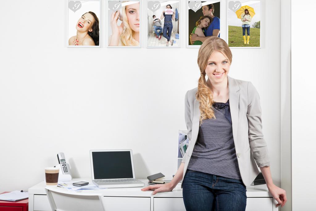 Ung kvinne i jeans og dressjakke står foran et arbeidsbord. På veggen bak kvinnen henger det fotografier av mennesker. Foto.