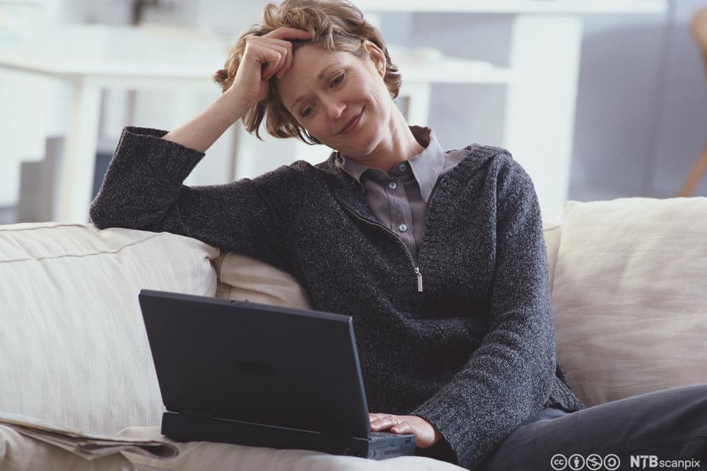 Kvinne som sitter i en sofa og leser på skjermen til en bærbar datamaskin. Foto.