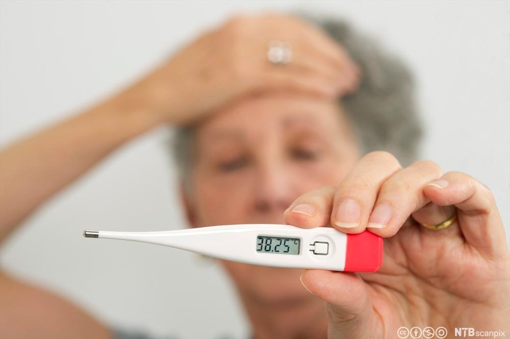 Kvinne holder seg på panna og viser fram et termometer som viser 38,25 grader. Foto.