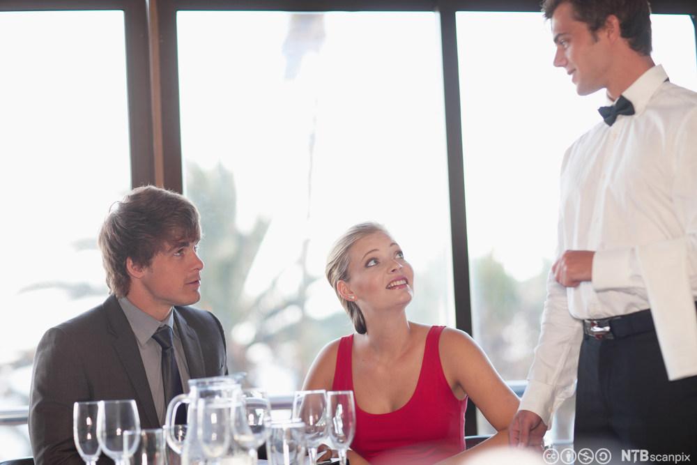 Ein servitør snakkar med to gjestar i restauranten. Foto.