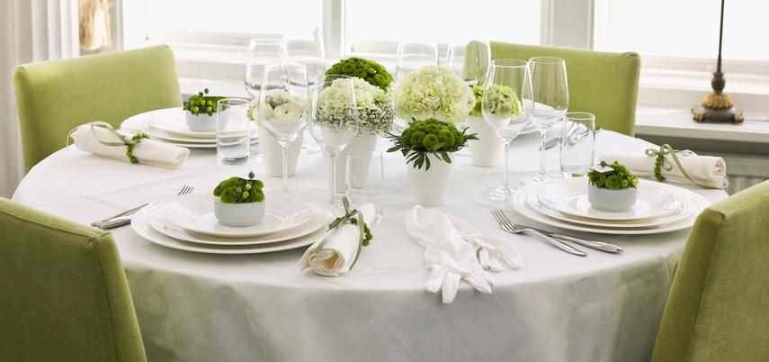 An elegantly set table with a white tablecloth. The chairs are green, as are the decorations on the table. There are white and green flowers. Photo.