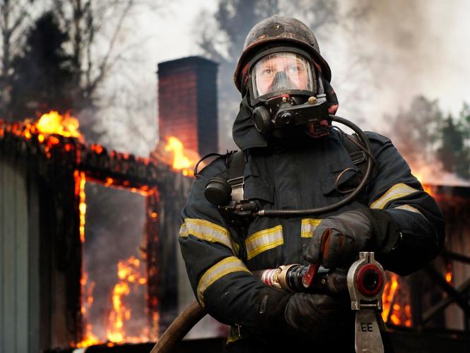 Foto av brannmann med vasslange framfor eit nedbrent hus.