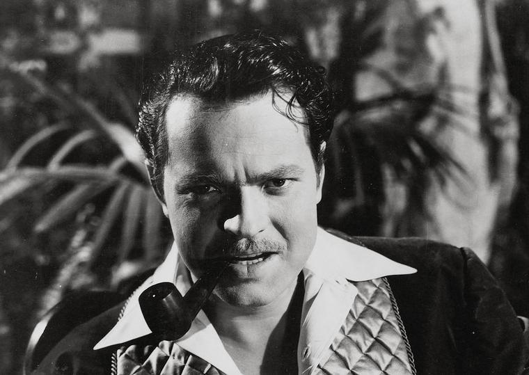 Portrett av skuespiller og regissør Orson Welles. Fotografiet viser en mann på rundt 40 år med tykt, relativt kort, svart hår som er gredd bakover. Han har ei pipe i munnen og et energisk ansiktsuttrykk.