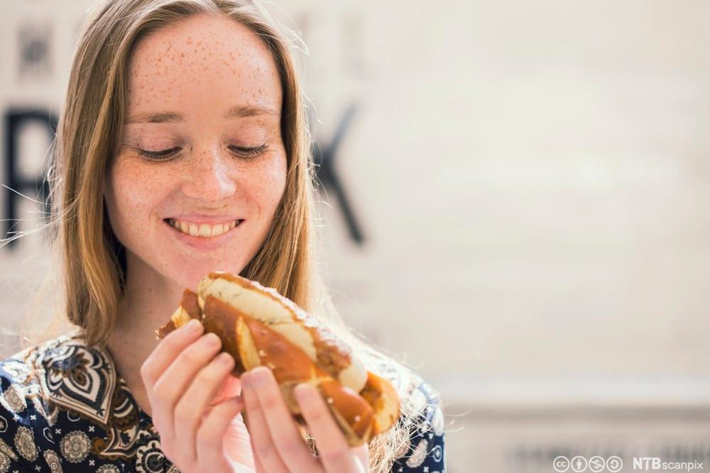 Ung kvinne spiser pølse i brød. Foto.