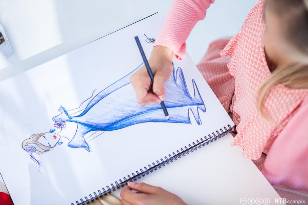 A girl is drawing a colorful sketch on a spiral-bound sketchbook. The sketch is of a woman with blonde hair, wearing a blue dress, who appears to be dancing or in motion. Photo.