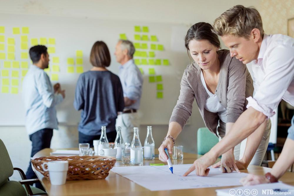 Folk i møteaktivitet: To personer står over et bord og peker på et ark, og tre personer står foran ei tavle med Post-it-lapper på. Foto.