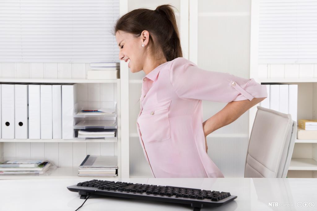 Kvinne ved kontorpult strekker seg med ryggsmerter. Foto.