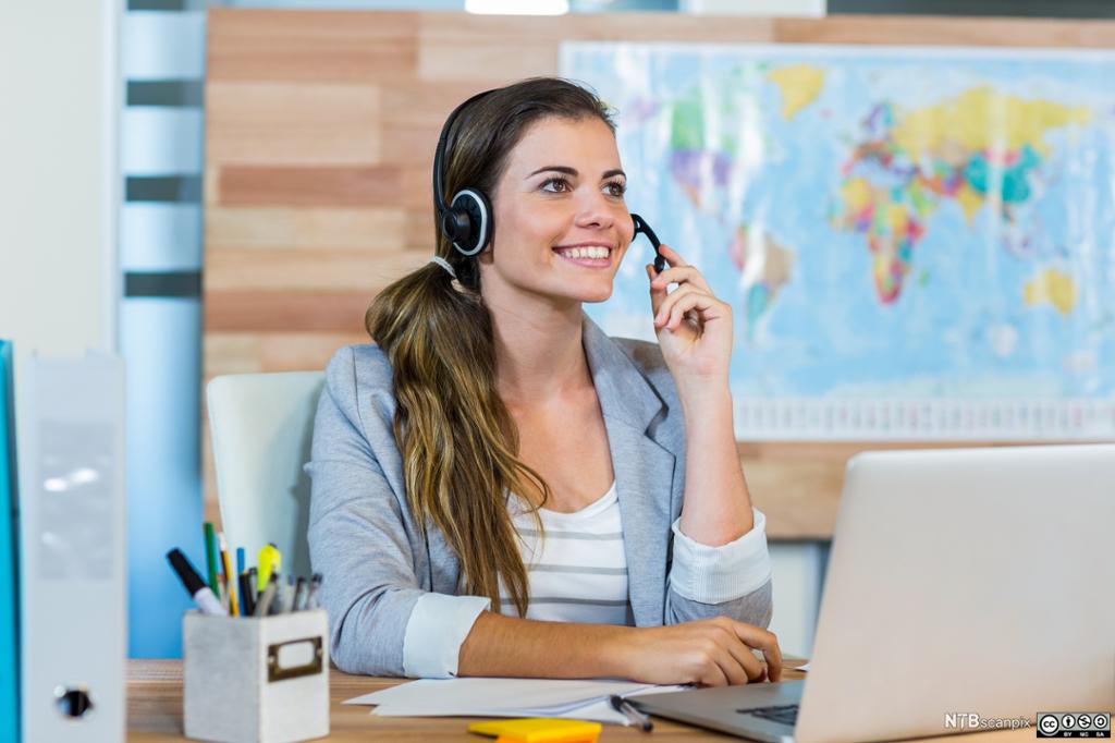 En smilende salgskonsulent ved et reisebyrå sitter med headset og PC. I bakgrunnen henger et verdenskart på veggen. Foto. 